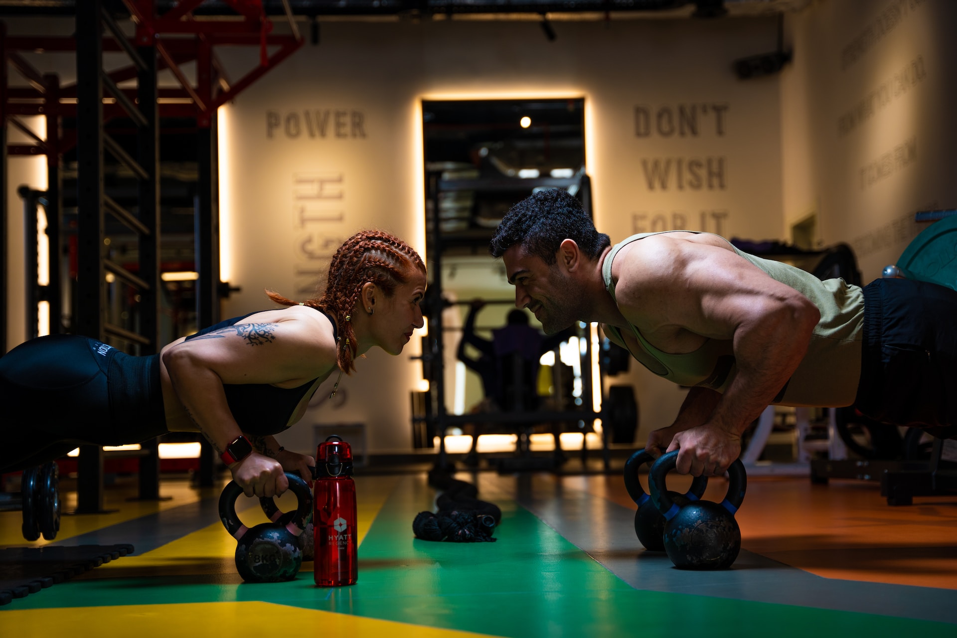 the image shows a female and a male athletes standing in the plank on the kettlebells